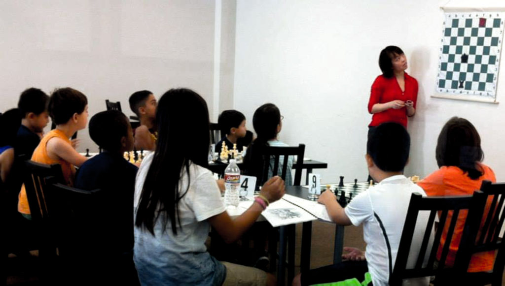 Kwunnie Ng (far right) providing valuable instruction to future Grand Masters at Chess 64 in Houston.  Kwunnie Ng is a Senior Tournament Director and FIDE National Arbiter.  She is the owner of The Chess Refinery, a pending 501(c)(3) in Houston.  Photo from the Chess 64 Facebook page.