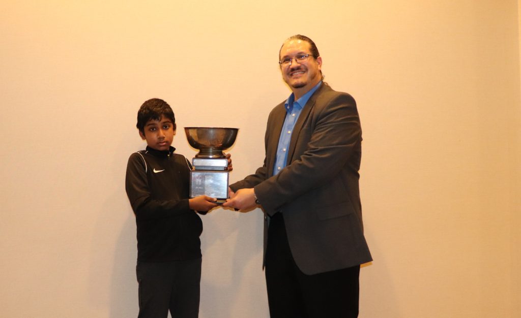 Chess Expert Vasee Nandhakumar (wearing the “Niki Swoosh”) receives his State Amateur Champion title and trophy from Chief Arbiter Louis Reed. Photo by Arlington Chess Club’s Jim Clarke.