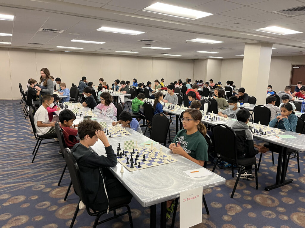 The Round Has Begun. Image of a lage room filled with chess players at their boards. By Caleb Brown.